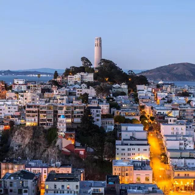 San Francisco's Coit Tower at dusk, with lighted streets before it and the San Francisco Bay behind it.